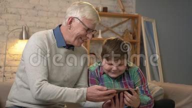祖父和孙子坐在沙发上，用平板电脑看着一部有趣的电影，笑着，微笑着。 家庭舒适